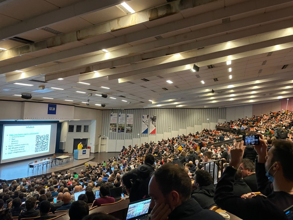 A picture of the opening keynote to FOSDEM 2023. The auditorium is full. On the podium there is a speaker, behind him a projected screen shows a slide with a QR code for the infodesk.